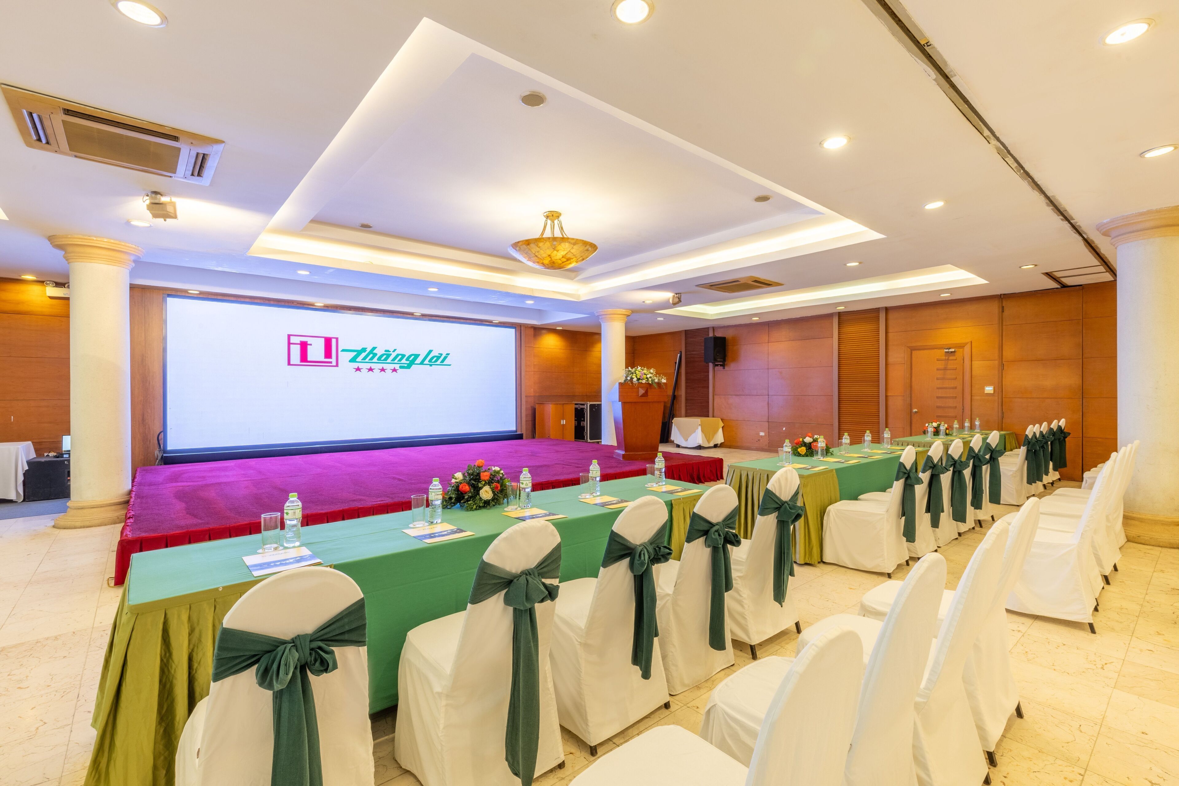 A conference room with a stage, a large screen displaying 'Thanglai', and rows of tables with chairs.