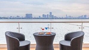 Ein Balkon mit Korbstühlen und einem Tisch, gedeckt mit Tellern und Getränken, mit Blick auf ein ruhiges Gewässer und eine Stadtlandschaft.