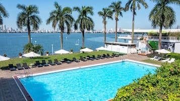 Una piscina con sillas de descanso y sombrillas, rodeada de palmeras y un horizonte urbano al fondo.