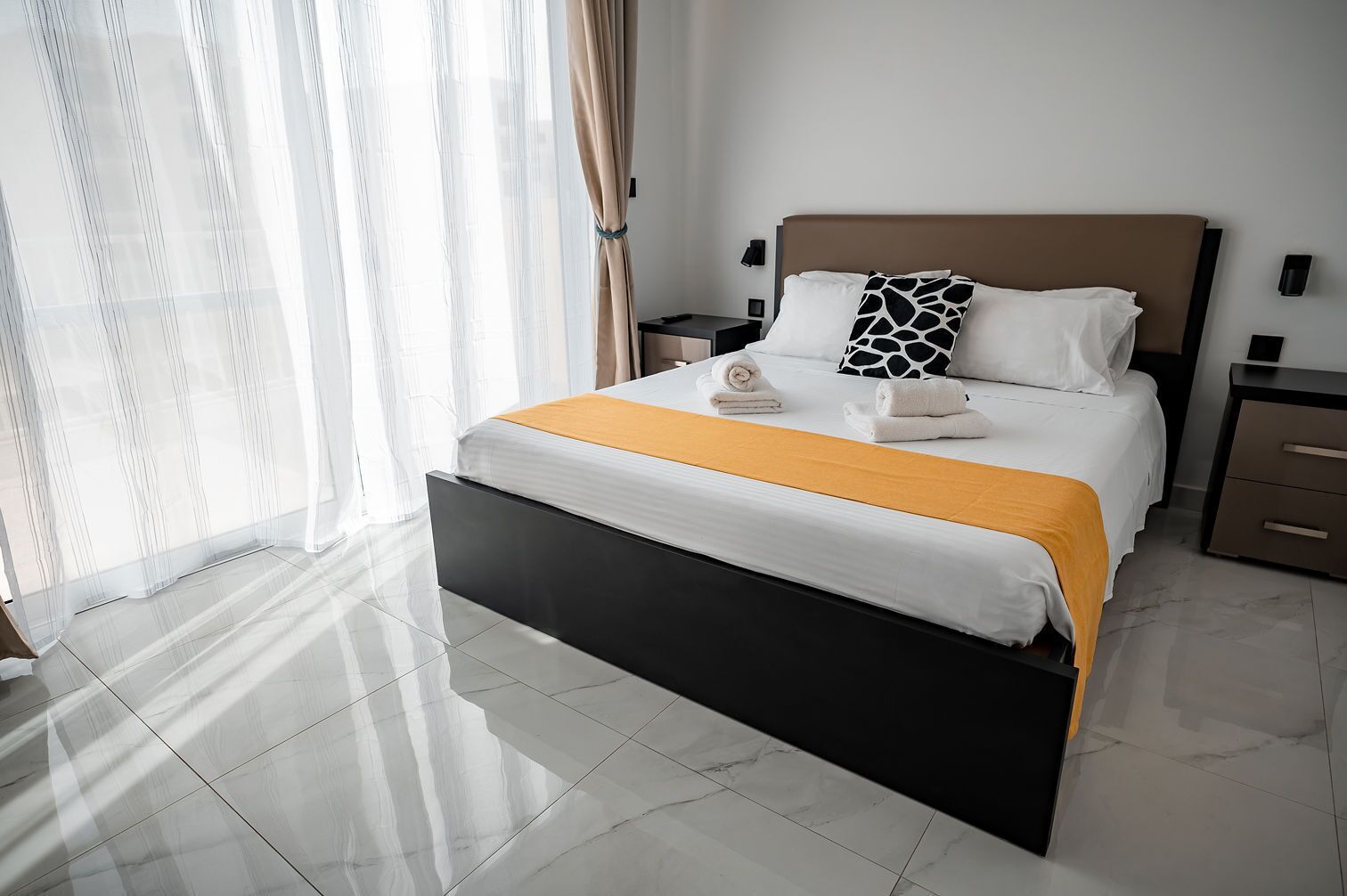 A modern hotel room with a large bed, a nightstand, a bedside table, and a window with sheer curtains.