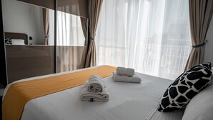 Een hotelkamer met een bed, een houten hoofdbord, een groot raam met dunne gordijnen en uitzicht op de stad.