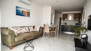 Een moderne woonkamer met een bank, een eettafel en stoelen. In de achtergrond is een keuken te zien met kasten en een spoelbak.
