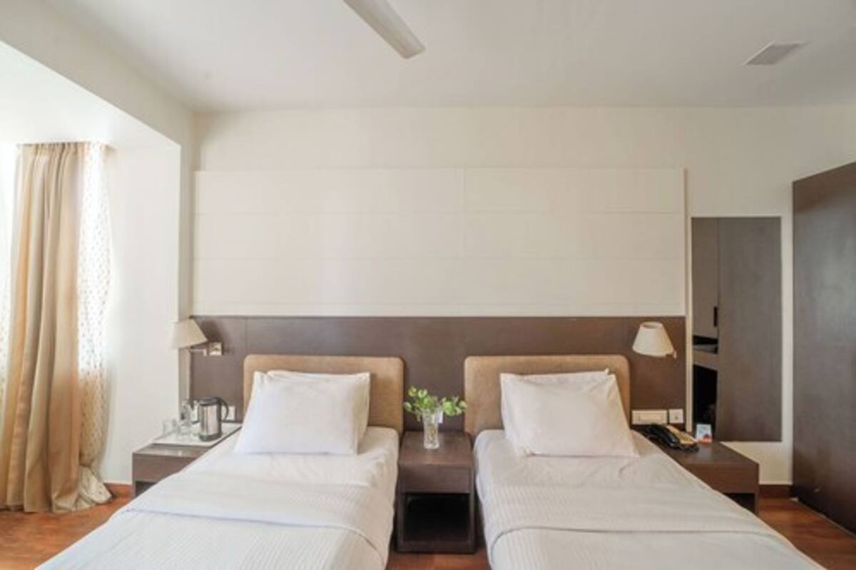 Ein Hotelzimmer mit zwei Betten, einem Nachttisch mit Lampe, einem kleinen Tisch mit Blumenvase und Blick auf die Stadt.