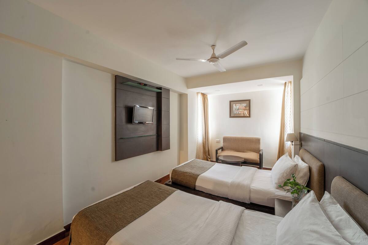 Ein Hotelzimmer mit zwei Betten, einem Deckenventilator, einem an der Wand montierten Fernseher, einem Sessel und einem Wandbild.