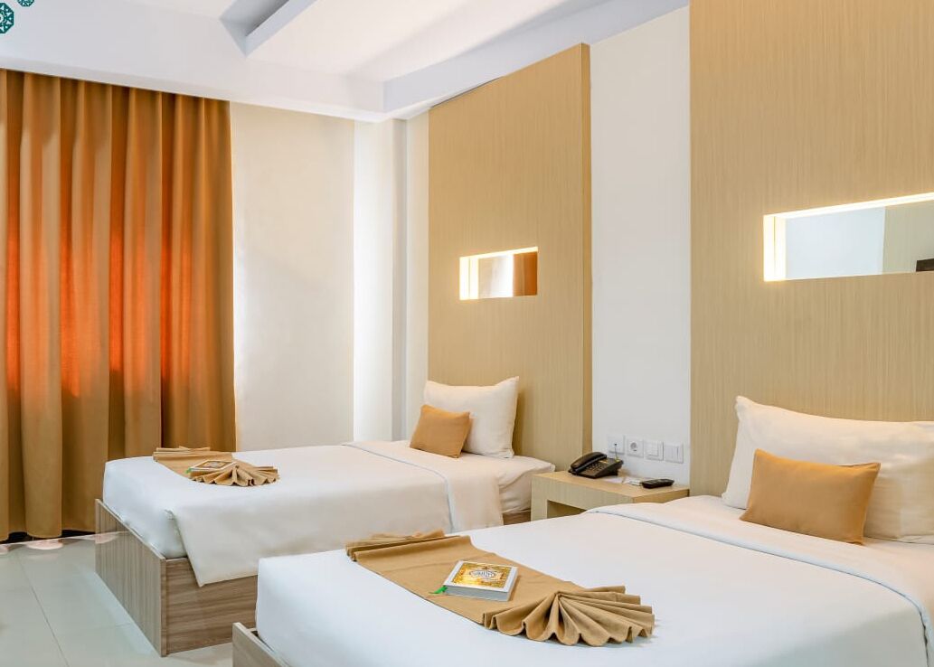 A hotel room with two beds, a wooden headboard, a bedside table with a phone, and a wall-mounted light fixture.