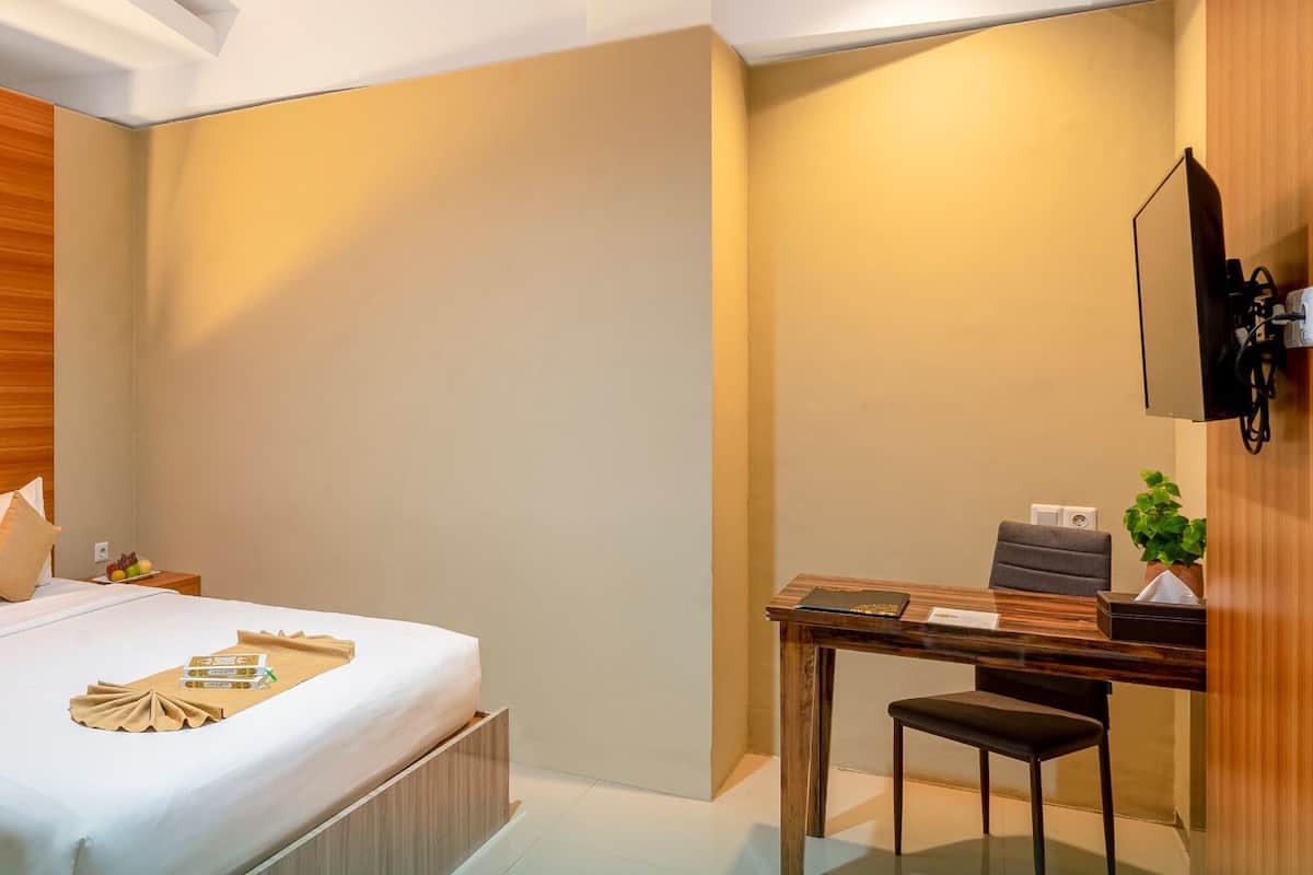 A hotel room with a wooden headboard, a bedside table with a phone and a lamp, a desk with a chair, and a small plant.