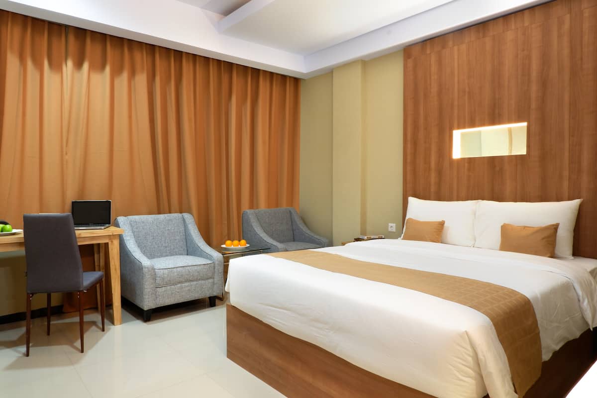 A hotel room with a large bed, a desk with a laptop, two armchairs, and a small table with fruit.