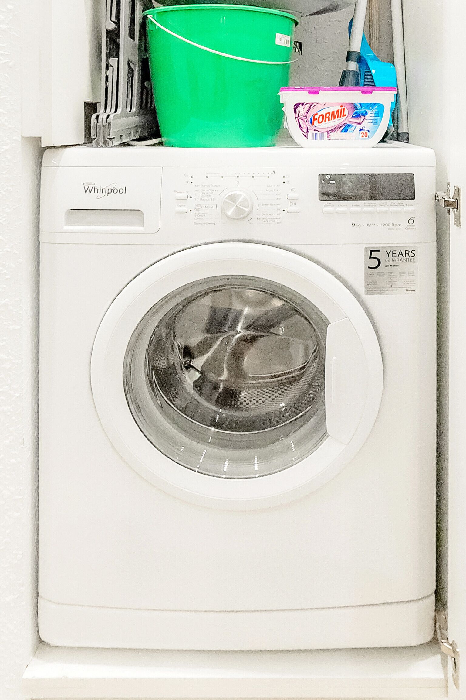 Een voorlader wasmachine van Whirlpool met een 5-jaren garantie sticker, een groene emmer en een fles Formil wasmiddel erboven.