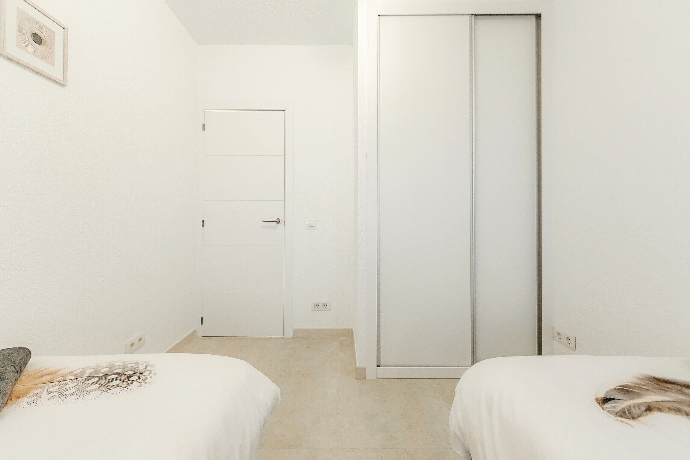 Ein schlicht eingerichtetes Hotelzimmer mit zwei Betten, einem Kleiderschrank und einer Tür zu einem weiteren Raum.