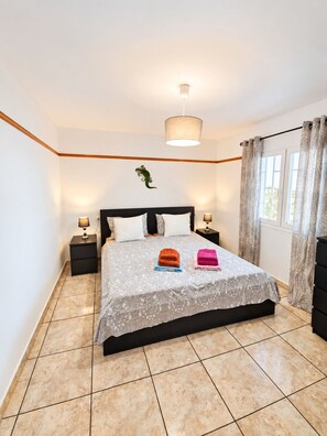 A bedroom with a bed, two bedside tables, a hanging light fixture, a window with curtains, and a wall-mounted plant. - Apartment Sea View  Holiday Valley -  Costa Adeje (Adeje)