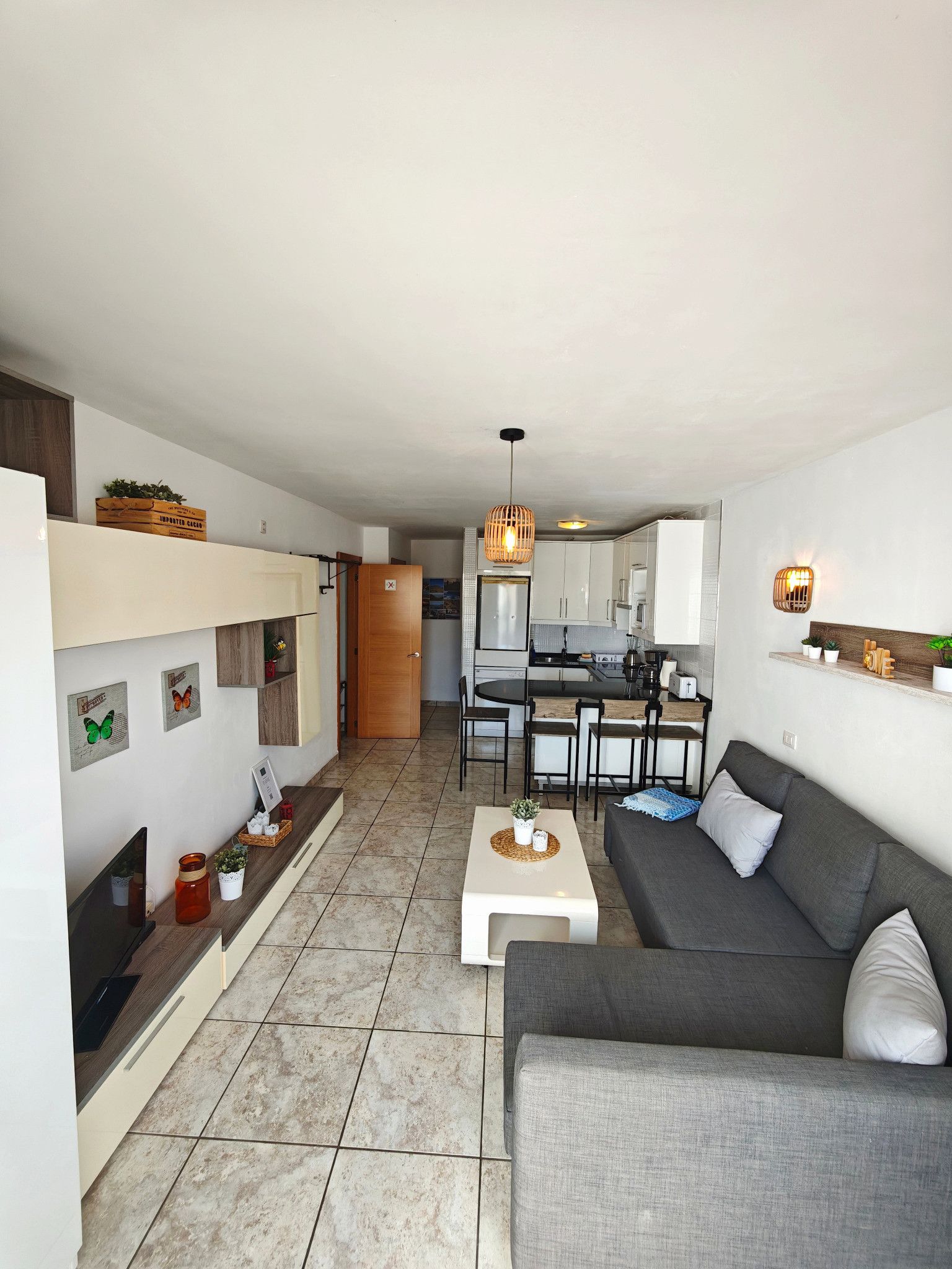 A modern living room with a grey sofa, a coffee table, and a TV. There is a kitchen in the background with white cabinets and a dining area.