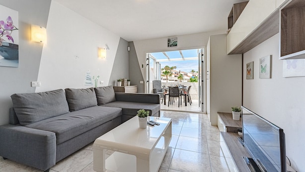 Una sala de estar moderna con un sofá gris, una mesa de centro blanca y un televisor. Hay un balcón con vistas a palmeras y edificios.