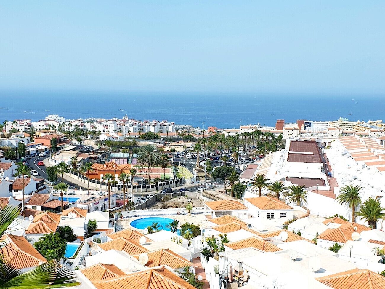 A coastal town with white buildings, a central pool, and palm trees.