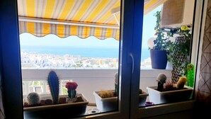 En balkon med udsigt over byen og havet, prydet med kaktusser i potter og en gulstribet markise.