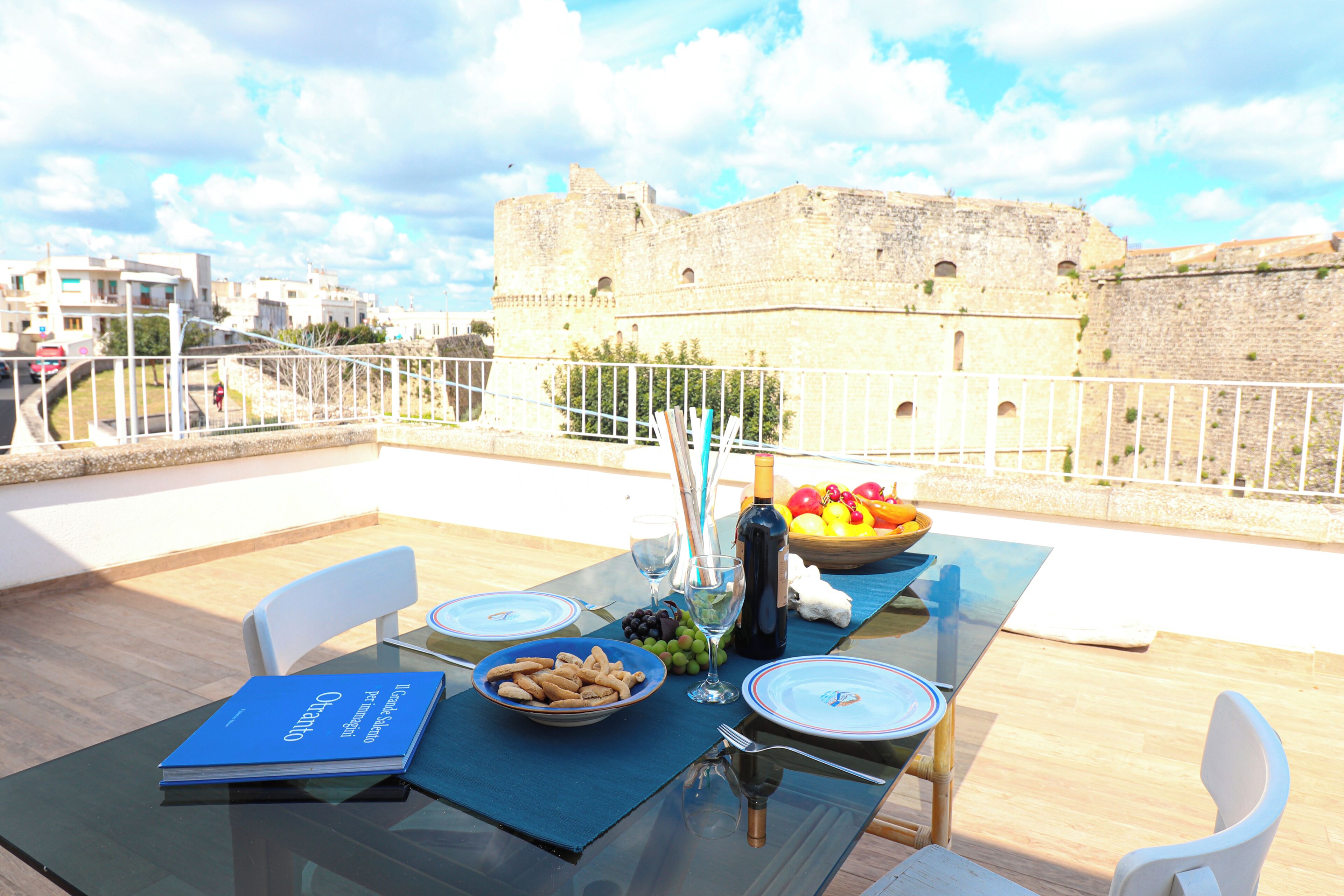 A table set for two with a book, a bowl of snacks, and a bottle of wine, overlooking a historic stone wall.