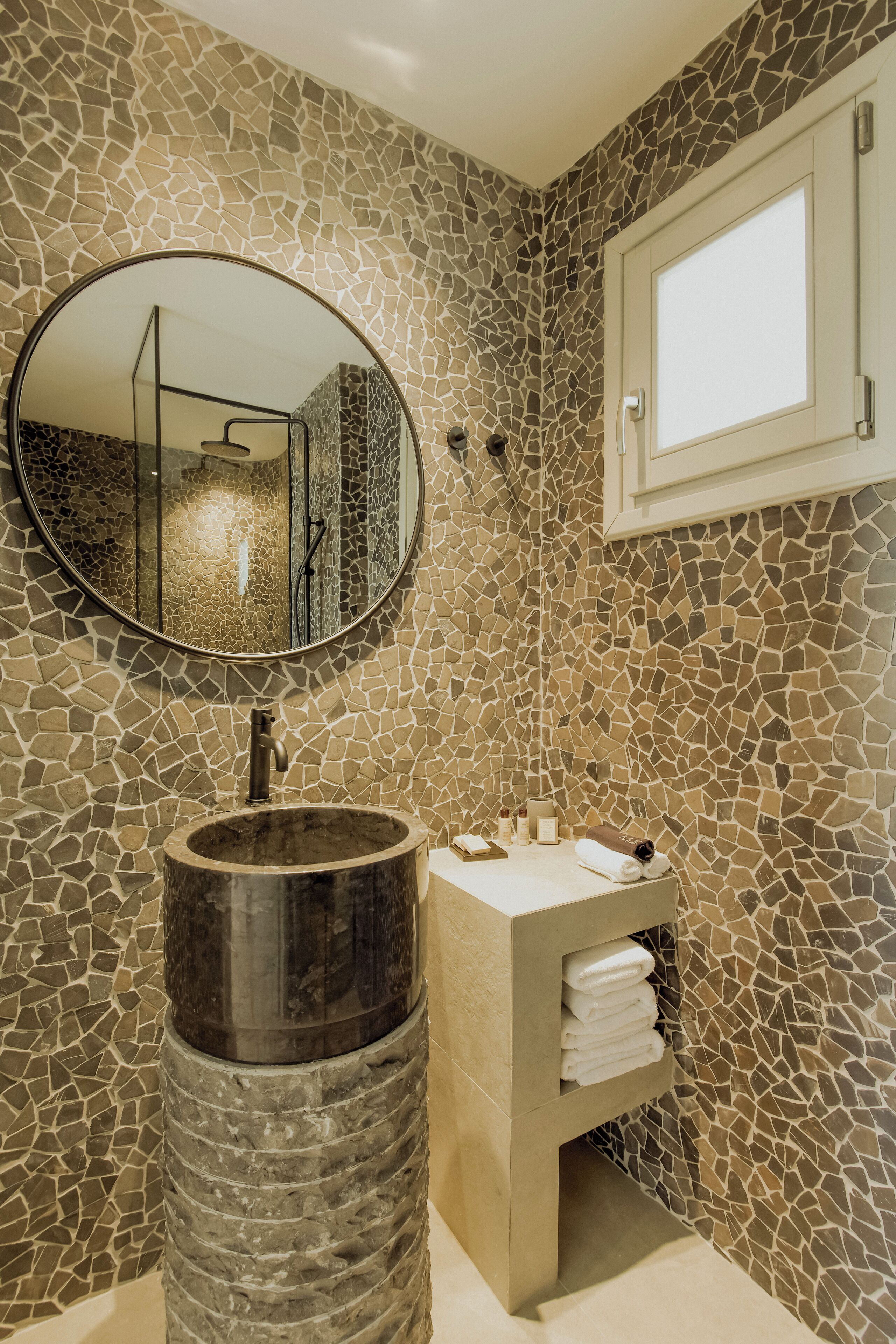 A bathroom with a mosaic wall, a round mirror, a wooden sink, and a towel rack.