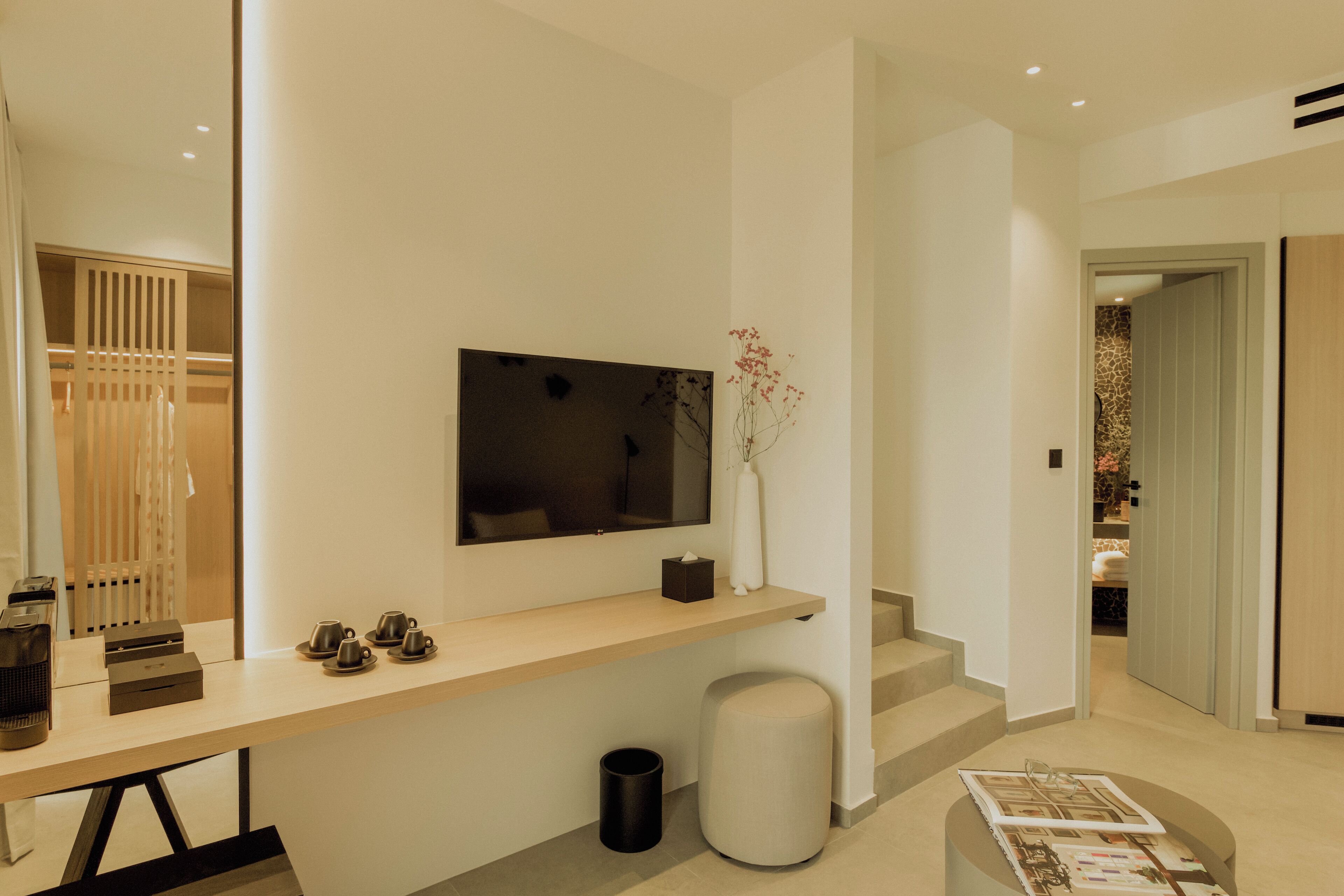 A modern hotel room with a minimalist design, featuring a long wooden desk with a flat-screen TV, a small vase with flowers, and a neatly arranged seating area with a bench and a small table.
