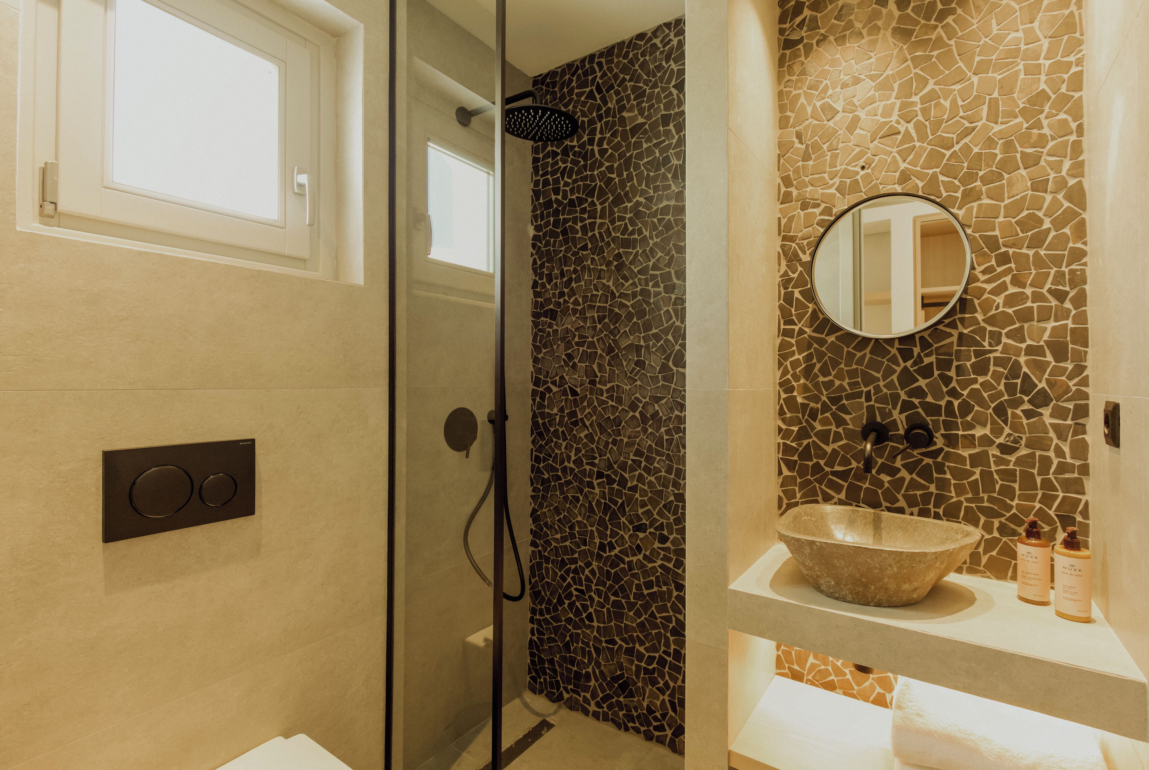 A bathroom with a glass-enclosed shower, a mosaic wall, a round mirror, and a stone sink.