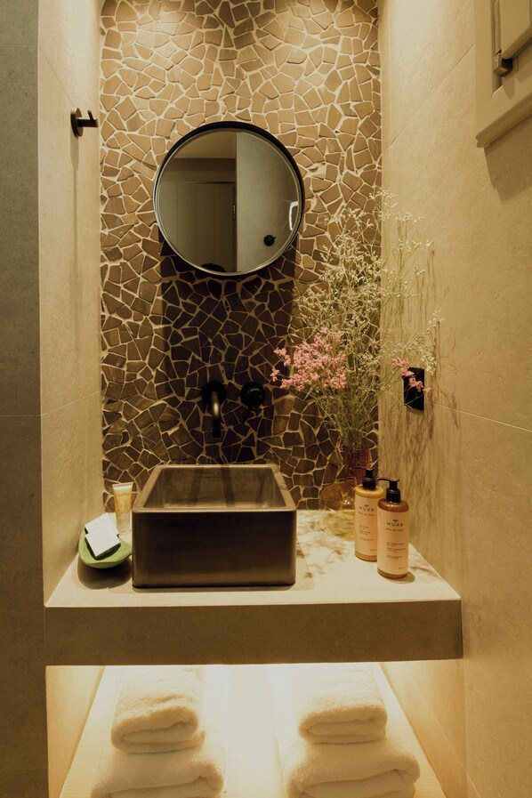 A bathroom with a stone mosaic wall, a round mirror, a sink, and a vase with flowers.