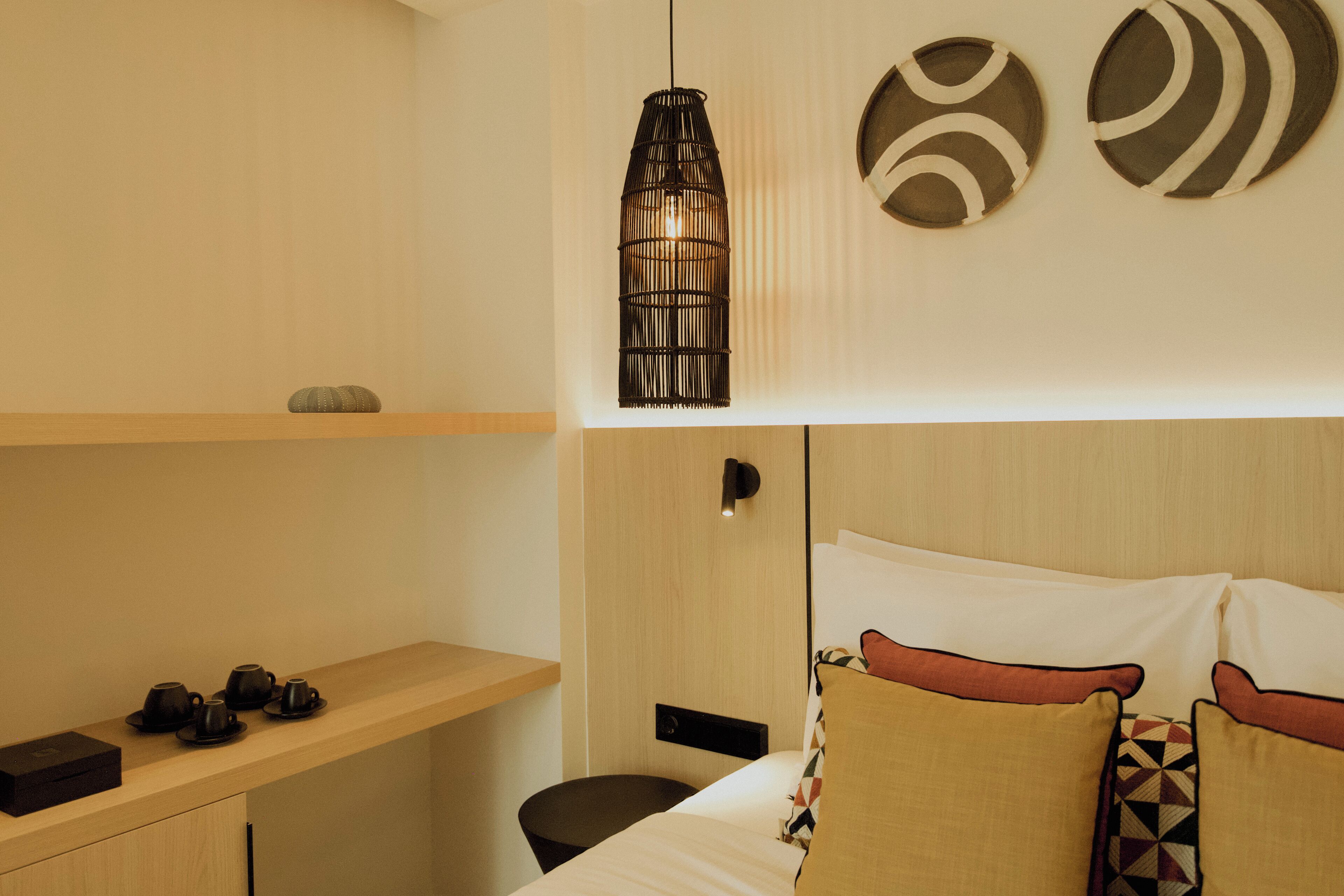A modern hotel room with a wooden desk, a bed with patterned pillows, a hanging light fixture, and wall art.