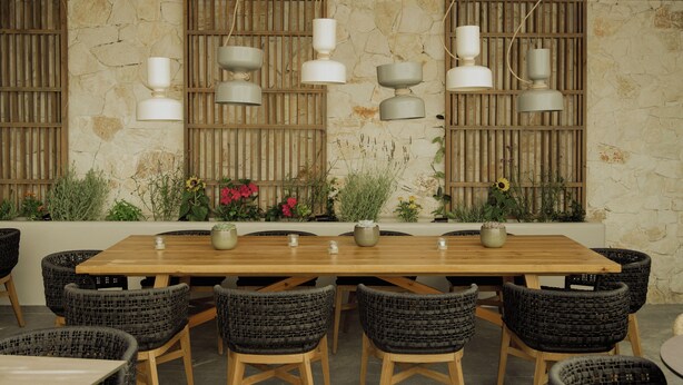 A wooden dining table with wicker chairs, hanging pendant lights, and a stone wall with vertical slats. - Opal Paros (Paros)