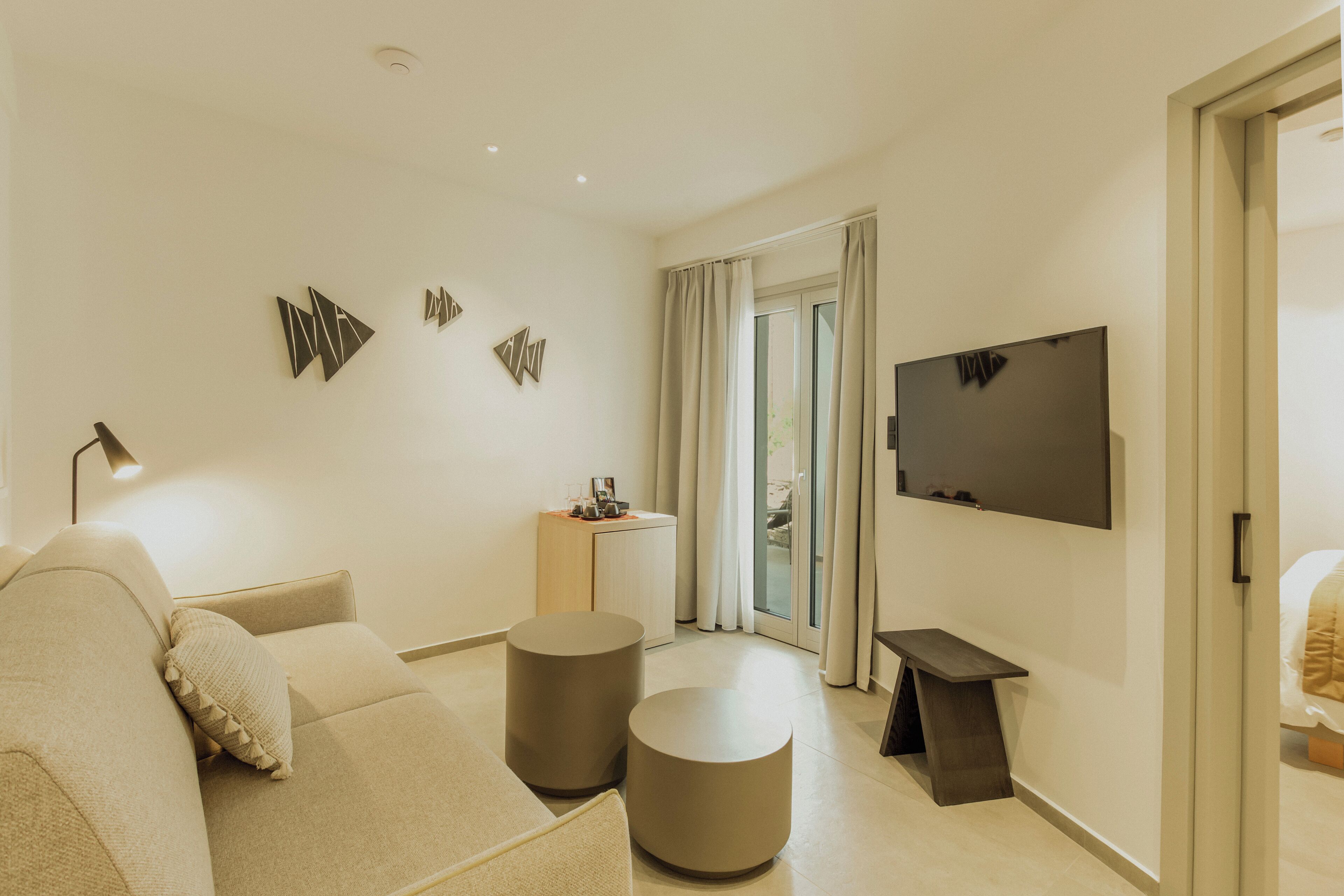A modern hotel room with a sofa, two round side tables, a flat-screen TV, and a desk with a lamp.