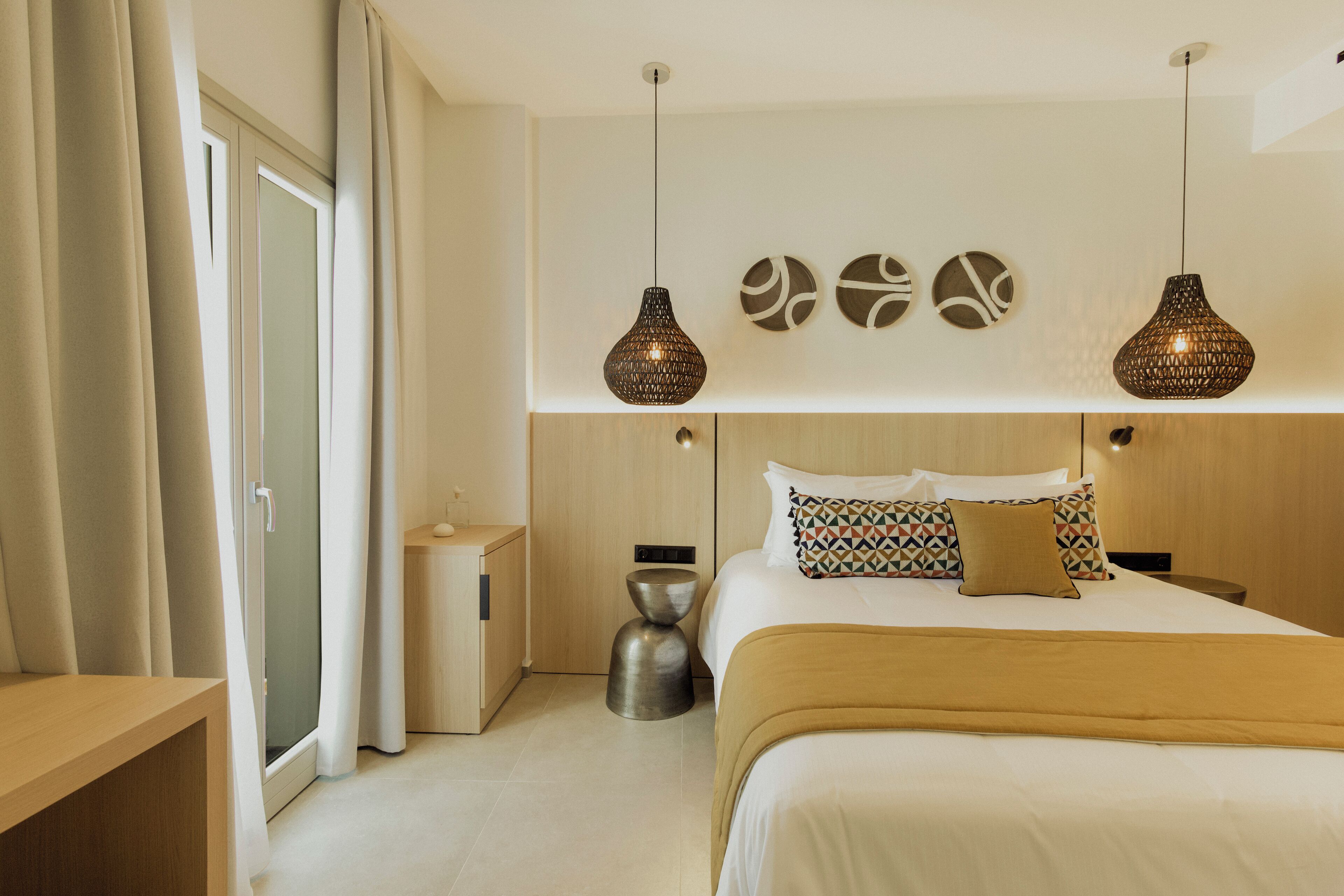 A modern hotel room with a large bed, two pendant lights, a wooden headboard, and a small side table.