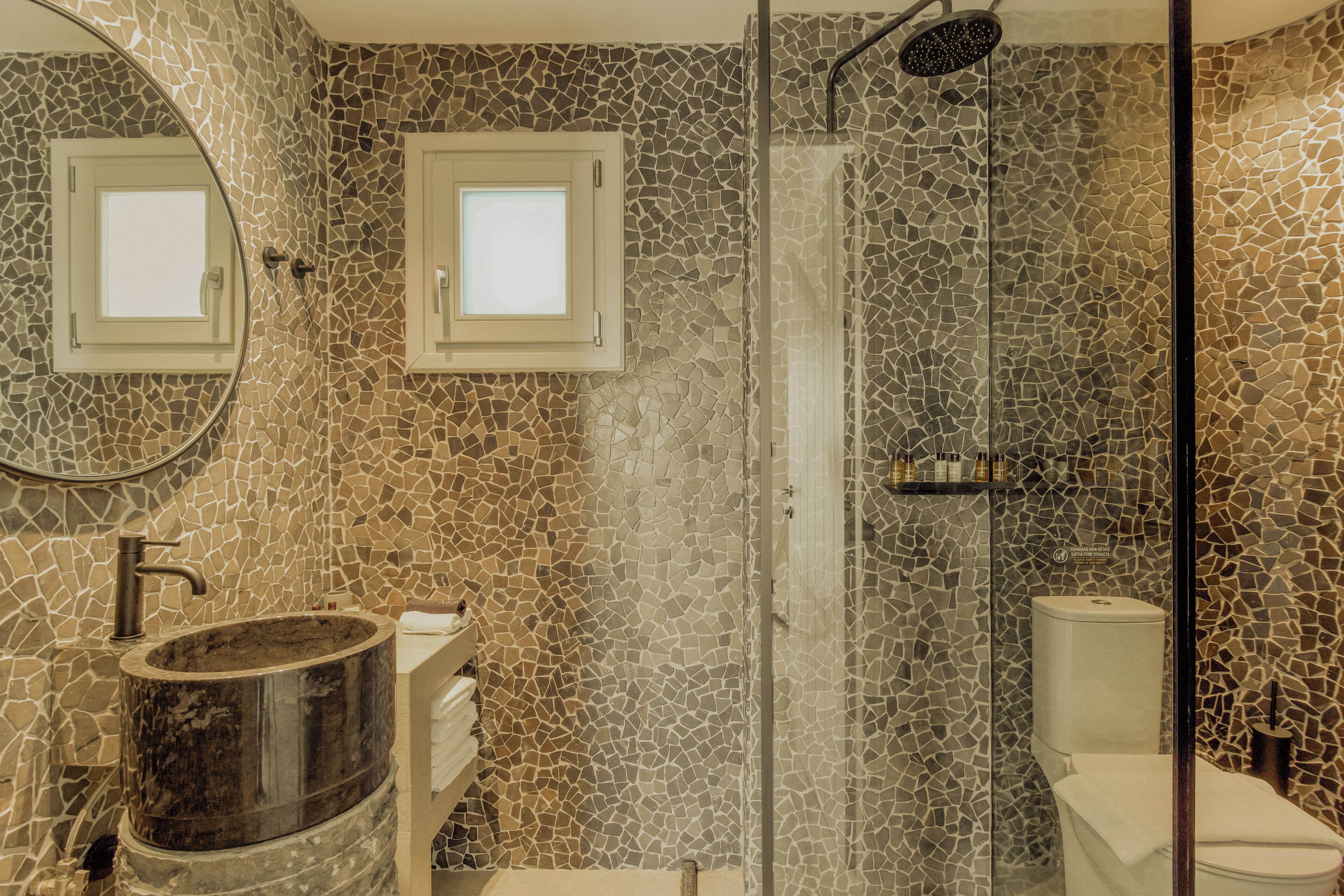 A bathroom with mosaic tile walls, a round sink, a glass-enclosed shower, and a toilet.