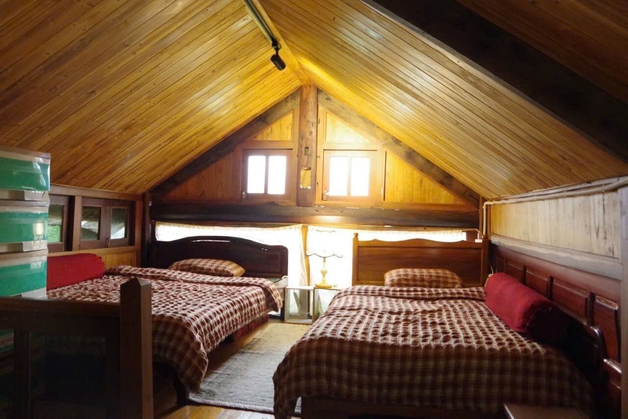 A cozy attic room with wooden beams, two beds with checkered bedding, a desk, and a window with curtains.