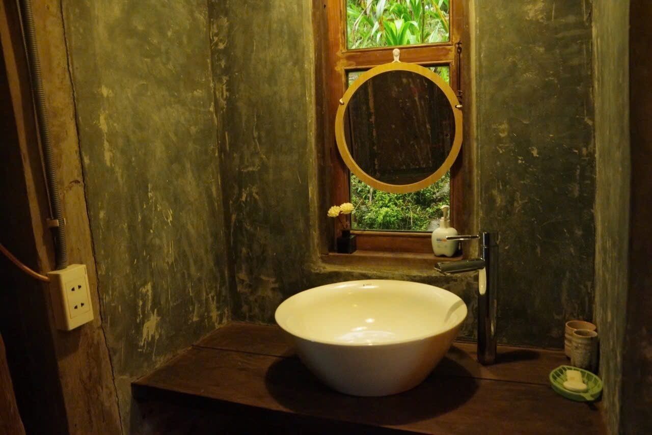 A rustic bathroom with a large white sink, a wooden-framed mirror, and a window with greenery visible outside.