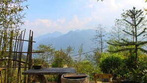 Un espace de restauration extérieur rustique, avec une table et un banc en bois, entouré de bambous et de verdure, et offrant une vue sur une chaîne de montagnes.