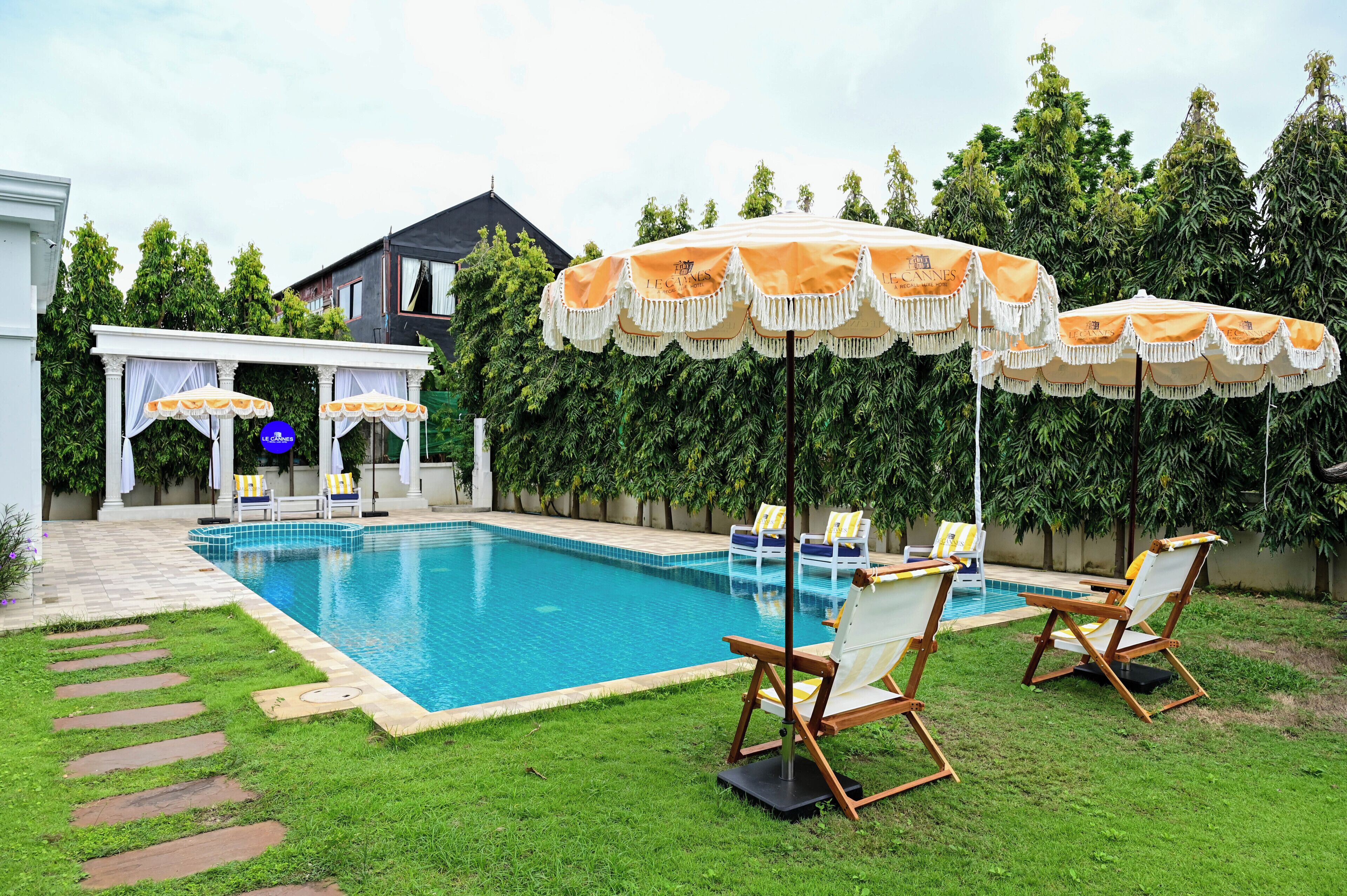 Ein Swimmingpool mit Liegestühlen und Sonnenschirmen, umgeben von viel Grün und einem Gebäude im Hintergrund.