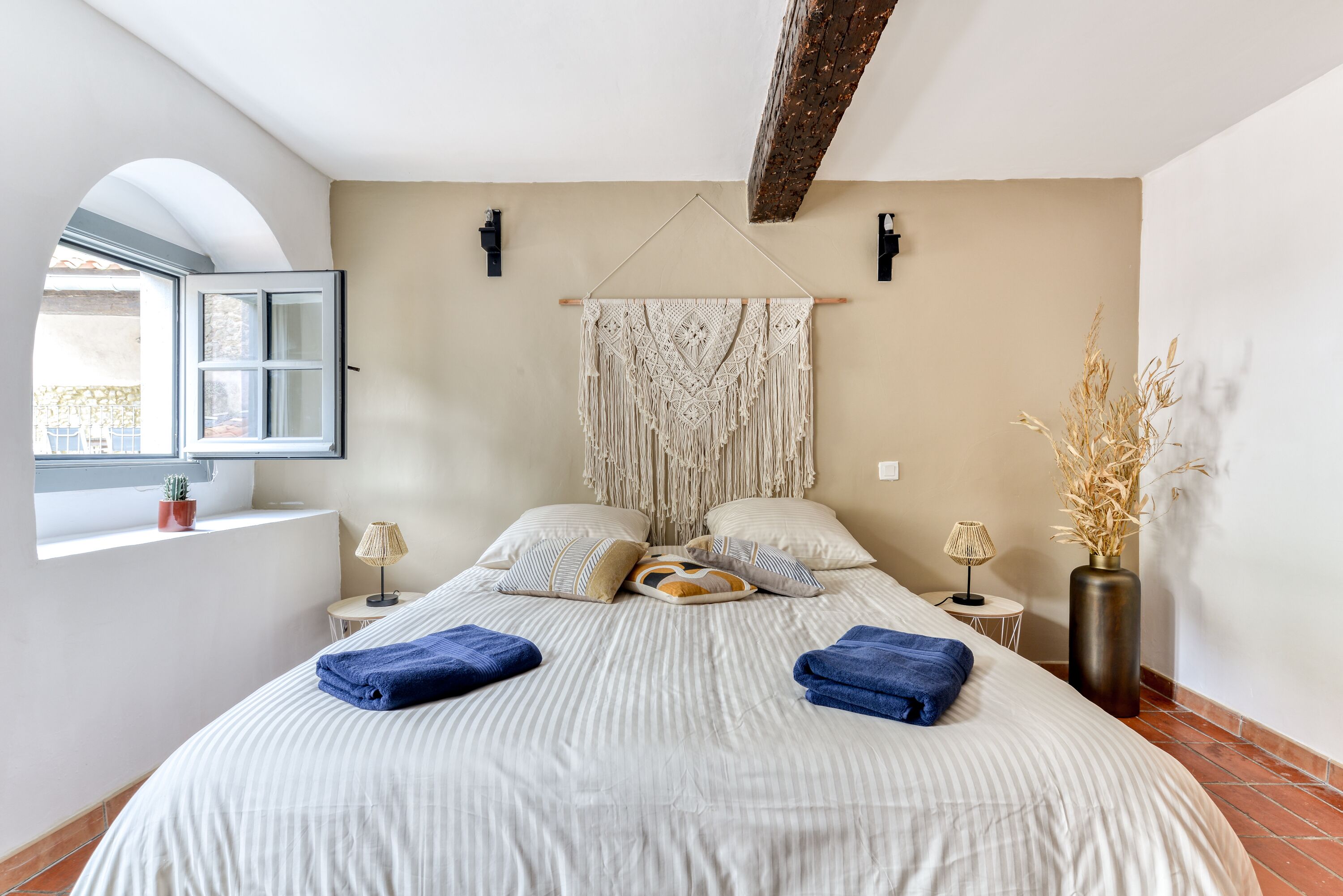 A bedroom with a bed, pillows, a macrame wall hanging, a window, a small table, and a vase with dried plants.