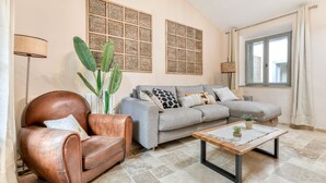 A living room with a leather armchair, grey sofa, and a coffee table. There are two wall-mounted decorative panels and a window with curtains.