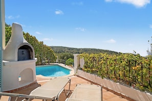 A poolside area with a barbecue grill, lounge chairs, and a view of the countryside.