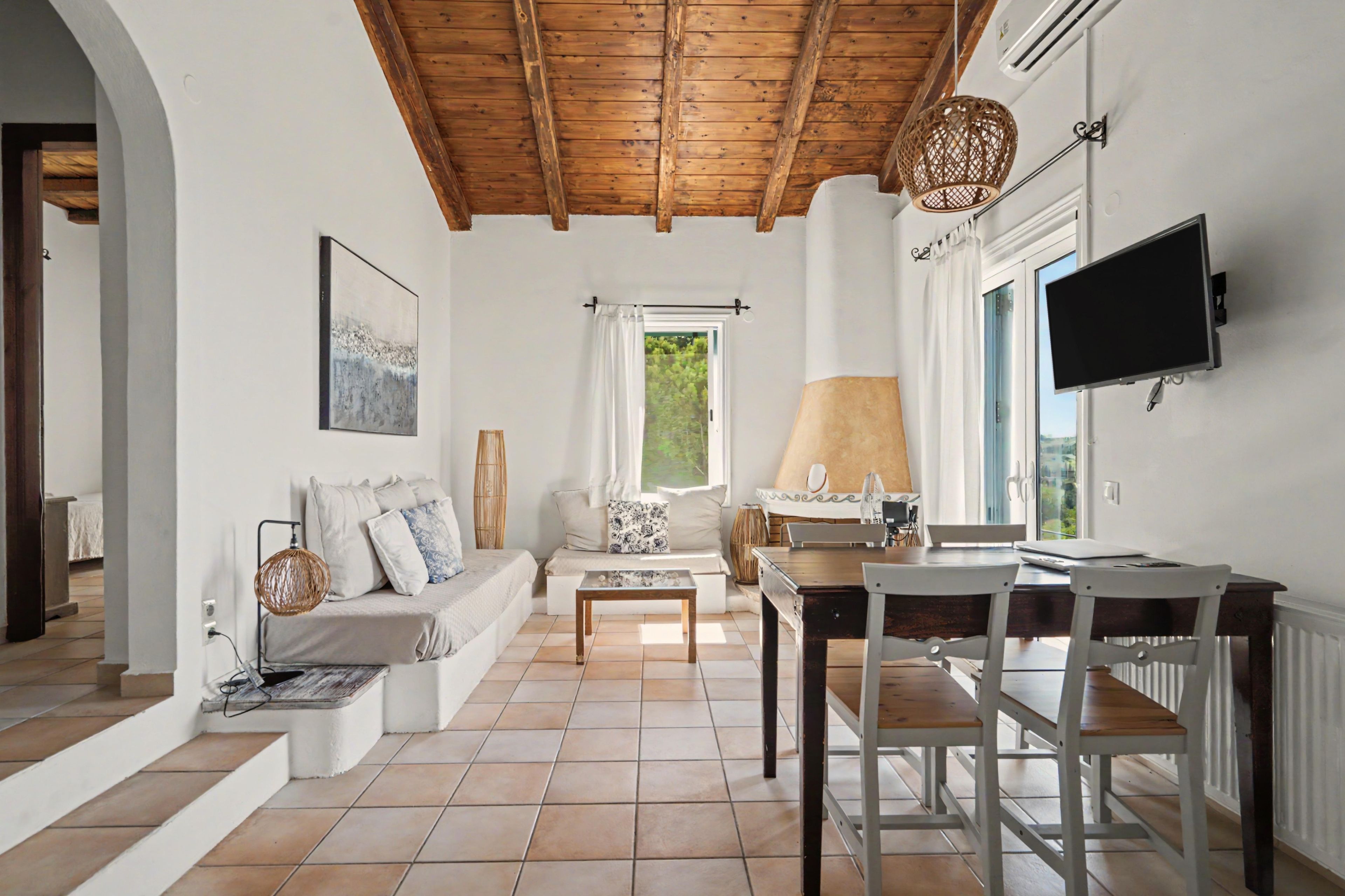 Un vaste espace de vie doté d’un plafond en bois, d’un ensemble de canapés, d’une table à manger avec des chaises, d’un téléviseur fixé au mur et d’une fenêtre donnant sur un paysage verdoyant.