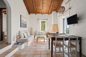 Een ruime woonkamer met een houten plafond, een zithoek, een eettafel met stoelen, een aan de muur bevestigde televisie en een raam met uitzicht op groen.