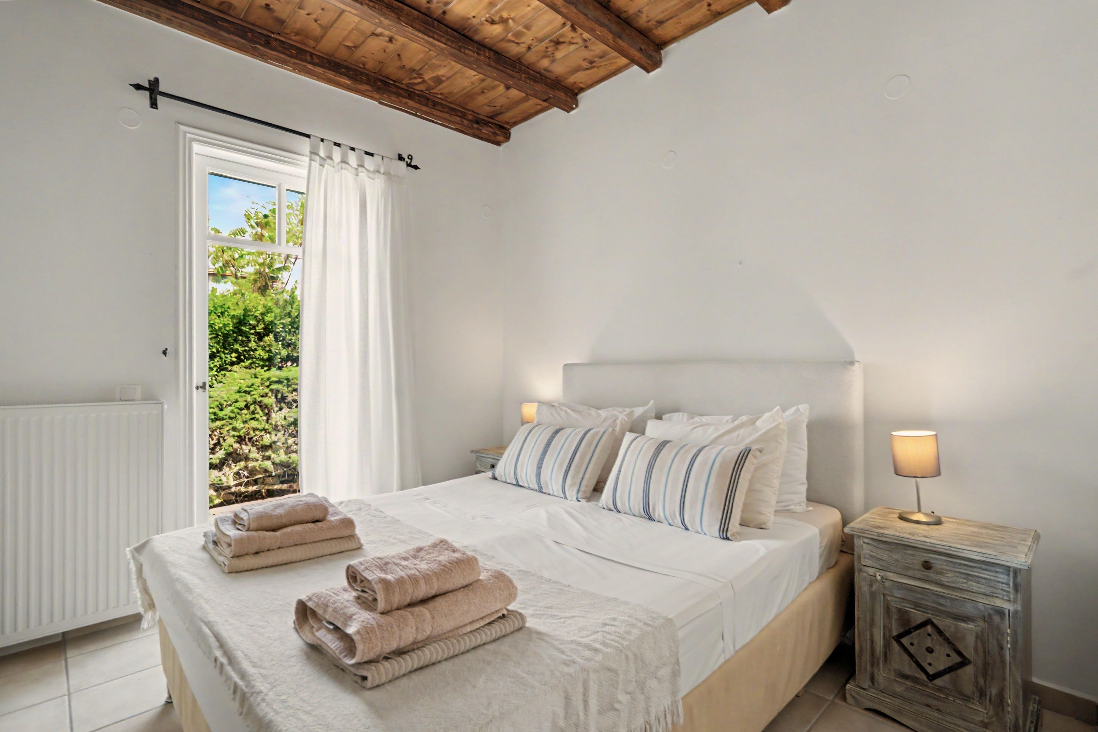 A bedroom with a bed, pillows, a bedside table with a lamp, a window with curtains, and a view of greenery outside.