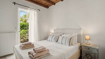 A bedroom with a bed, pillows, a bedside table with a lamp, a window with curtains, and a view of greenery outside.