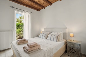 A bedroom with a bed, pillows, a bedside table with a lamp, a window with curtains, and a view of greenery outside. - Holiday Home 'Villa Ostria' with Sea View, Private Pool and Wi-Fi (Tsoukalades)