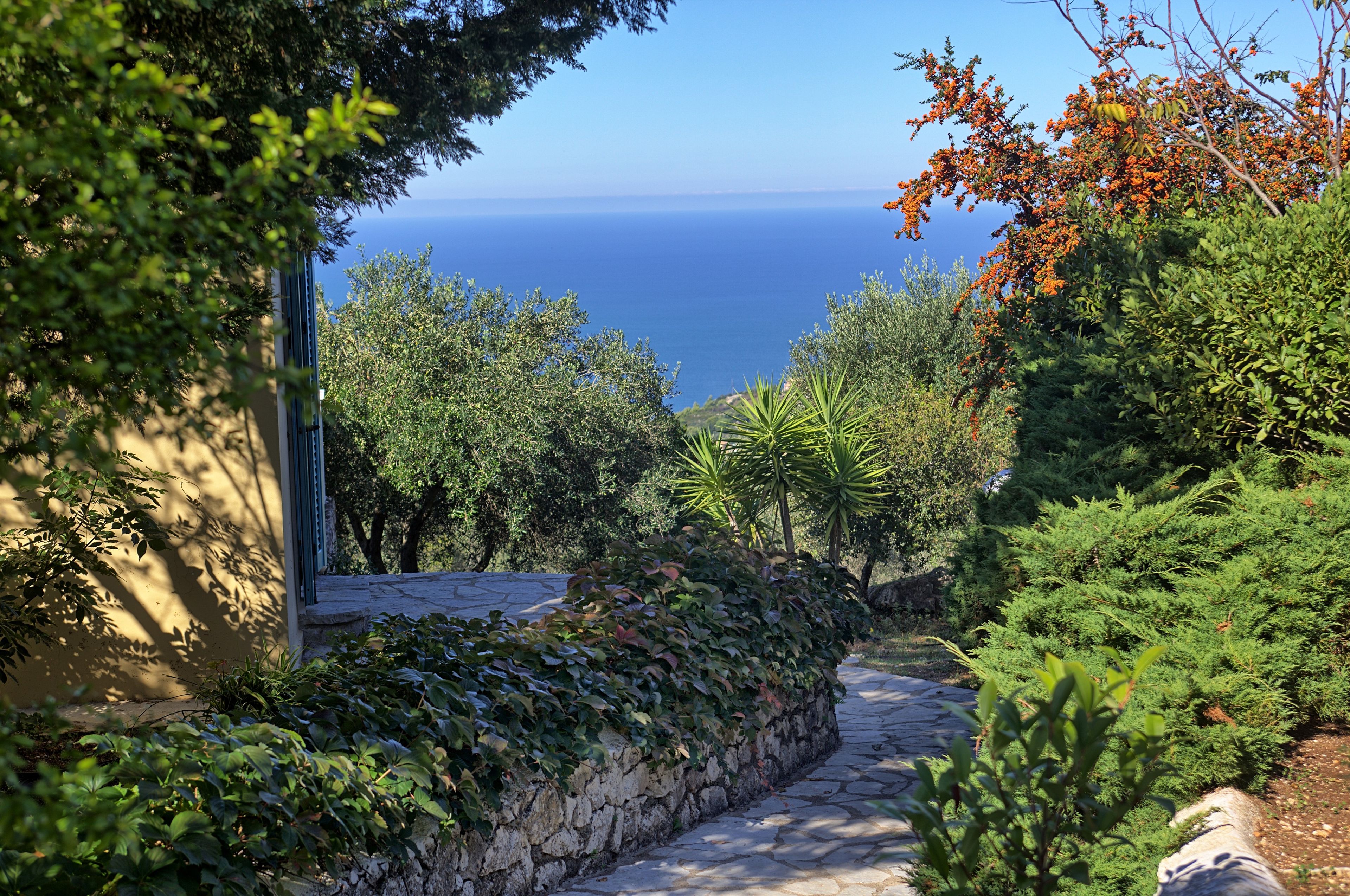 Un sentier de pierres menant à un point de vue sur la mer, bordé d’une végétation luxuriante et délimité par un mur de pierres.