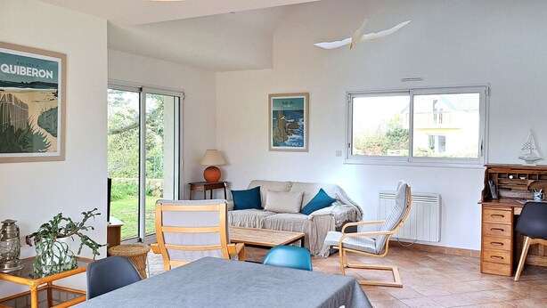 Una habitación bien iluminada con una mesa de comedor, sillas, un sofá y un escritorio. Amplias ventanas dejan entrar luz natural y ofrecen vistas del exterior.