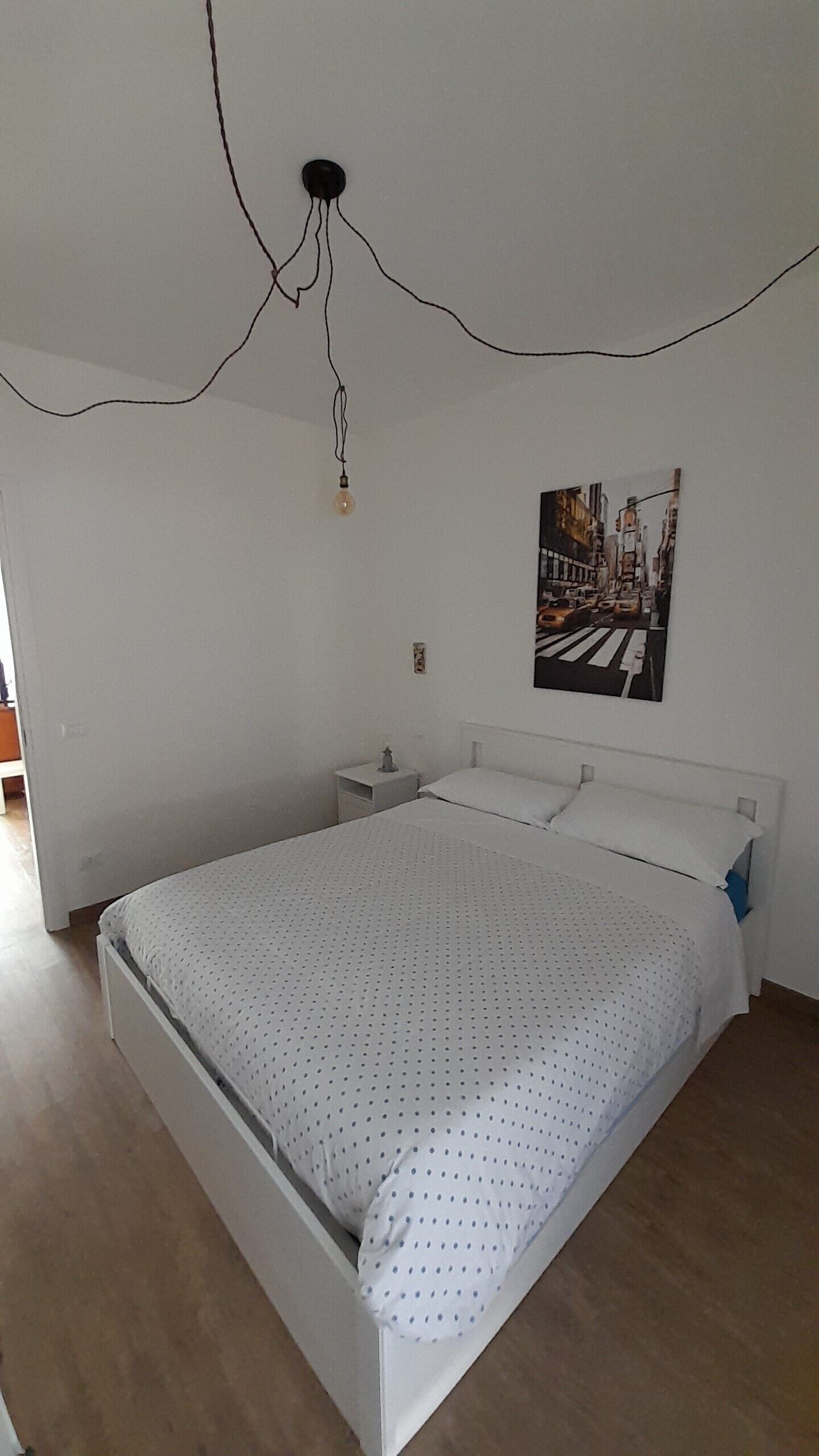 Quarto com cama, mesa de cabeceira, luminária na parede e um pôster.