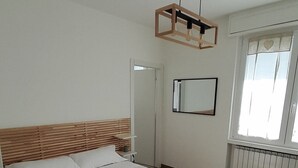 Un dormitorio con estructura de cama de madera, una mesita de noche, una lámpara colgante, una ventana con cortinas y un radiador.