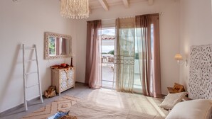Una habitación con techo de madera, un candelabro, un espejo grande, un armario de madera, una cama con cabecera estampada y vistas por una puerta de cristal.