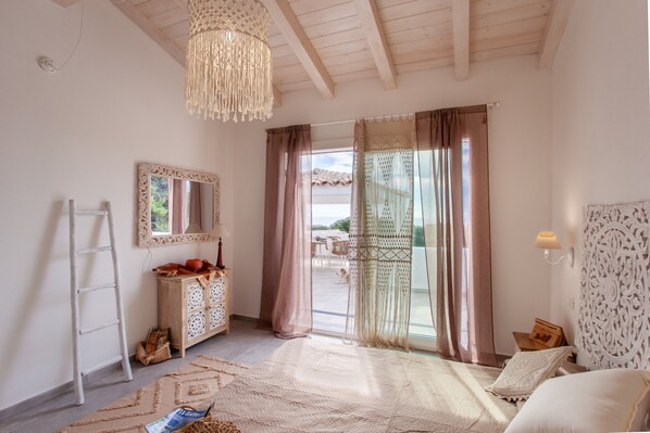 A room with a wooden ceiling, a chandelier, a large mirror, a wooden cabinet, a bed with a patterned headboard, and a view through a glass door.