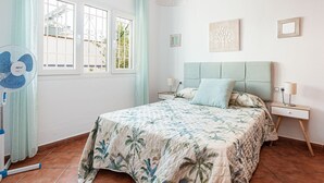 Een slaapkamer met een bed, nachtkastjes, een ventilator en een raam met gordijnen.