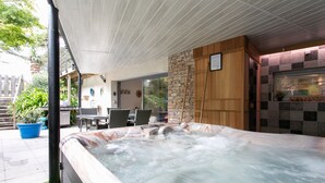 Un jacuzzi con pared de piedra y paneles de madera, un patio cubierto con mobiliario exterior, y vista a un jardín.