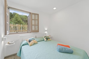 A bedroom with a bed, pillows, a nightstand, a window with a view of a fence and trees, and a wall-mounted light. - Holiday Home 'Ikigai' with Mountain View, Garden & Wi-Fi (Buggerru)