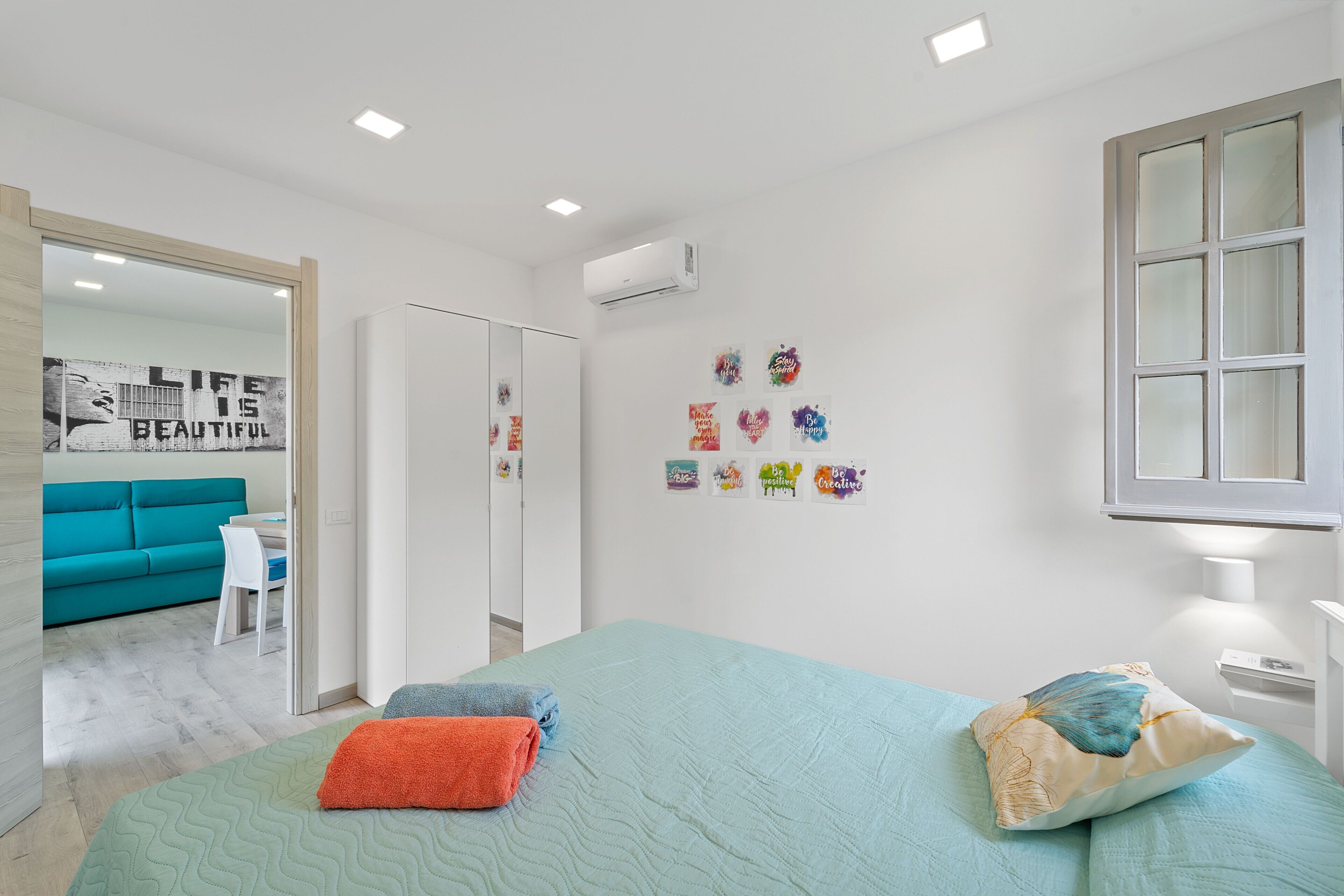 A modern bedroom with a bed, pillows, a wall with motivational stickers, a window, and a door leading to another room.